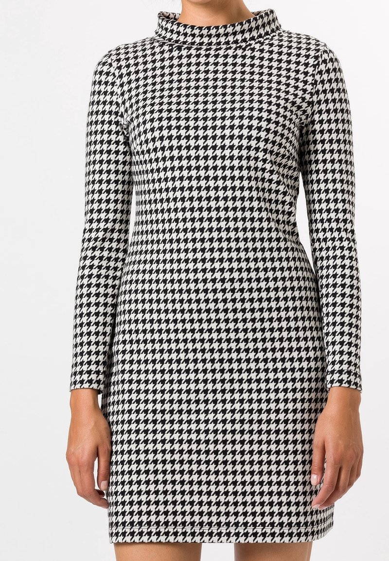 Personne portant une robe à manches longues à motif pied-de-poule noir et blanc avec un col montant, debout devant un fond uni.