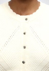 Gros plan d'un cardigan tricoté crème avec un col festonné et quatre boutons en argent en forme de cœur sur le devant.