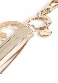 Gouden metalen sleutelhanger met een getextureerde ronde charme, een tassel van glad materiaal en een kleine ronde tag met de inscriptie "MIRIADA."