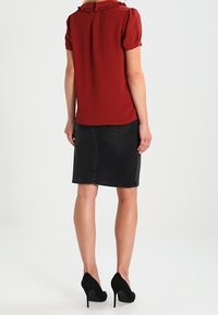 Blouse rouge avec un col à volant, manches courtes bouffantes, associée à une jupe en denim noir et des chaussures à talons hauts noires. Fond neutre.