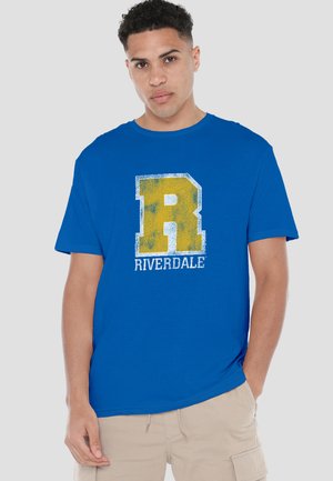 Young man wearing a blue T-shirt with a large yellow "R" and "RIVERDALE" text, paired with beige cargo shorts, standing against a plain background.