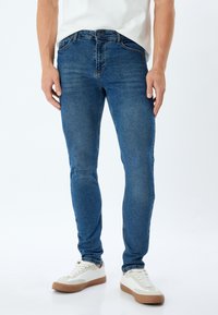 Vaqueros ajustados de mezclilla azul, con cinco bolsillos, una cintura con trabillas para cinturón y una leve textura desgastada en la tela.