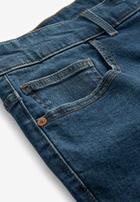Close-up van een blauwe denim jeanszak met zichtbare stiksels, een klinknagel en een riemlus op een witte achtergrond.