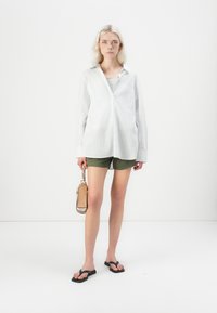 Camicia bianca con strisce azzurro chiaro, pantaloni corti verde oliva, ciabatte nere e una piccola borsa beige con tracolla intrecciata.