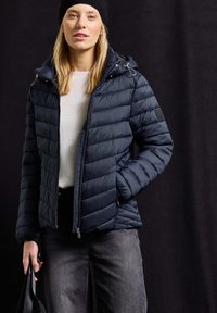 Doudoune marine avec fermeture éclair sur le devant, motif en losanges et capuche. Un bonnet noir en tricot, une chemise gris clair et un jean foncé complètent la tenue.