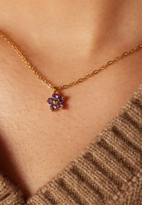 Gouden ketting met een bloemvormige hanger met vijf paarse edelstenen, geplaatst tegen een achtergrond van gestructureerde huid en bruin gebreide stof.
