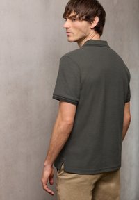 Jeune homme aux cheveux bruns courts portant un polo gris foncé et un pantalon beige, légèrement tourné sur le côté contre un mur uni.