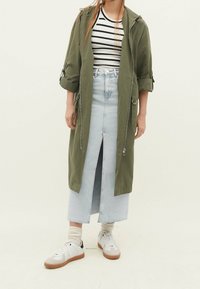 Olive green hooded long jacket over a black and white striped top, paired with a light blue denim skirt featuring a front slit and white sneakers.