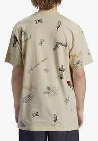 Camiseta de algodón beige con varios estampados gráficos en negro y colores, que incluyen dados, una cruz, telarañas y llamas a lo largo de la tela.