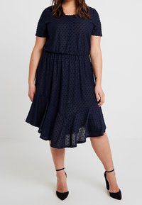 Mörkblå klänning med kort ärm och texturerat prickmönster, elastisk midja och volangnederdel, i kombination med svarta högklackade skor.