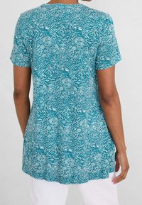 Teal kortärmad tunika med ett vitt blommönster och vågigt mönster, med en avslappnad passform och rundad nederkant. Tyget verkar mjukt och lätt.