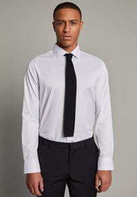 Camisa blanca de manga larga con un cuello estructurado, combinada con una corbata negra texturizada. Pantalones oscuros de corte formal completan el atuendo.