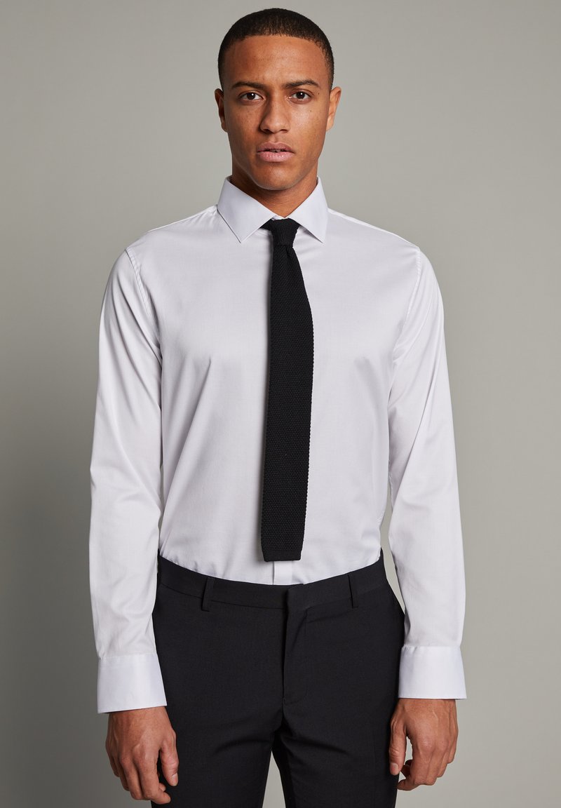 Camisa blanca de manga larga con un cuello estructurado, combinada con una corbata negra texturizada. Pantalones oscuros de corte formal completan el atuendo.