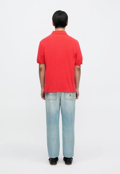 Polo rouge à col classique, associé à un jean denim bleu clair. Le jean présente une légère décoloration et un patch de logo vert à l'arrière.