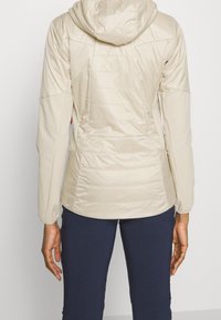 Häilyvy beige pehmustettu takki hupulla, jossa on sileä rakenne, kontrastiväriset saumat ja istuva muotoilu. Takakatsaus joustavilla hihoilla.