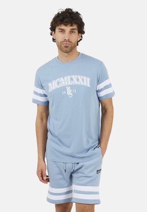 Man met krullend haar draagt lichtblauwe korte broek en T-shirtset met witte strepen en Romeinse cijfers op het shirt, staand met één hand in de zak.