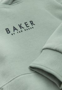 Jasnozielona tkanina z czarnym tekstem o treści "BAKER by Ted Baker" oraz widoczny złożony mankiet długiego rękawa.