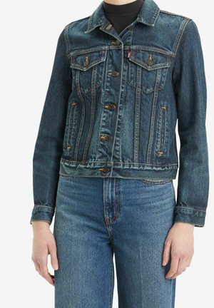 Osoba nosząca niebieską kurtkę dżinsową z zapięciem na guziki oraz dopasowane wysoko podniesione niebieskie dżinsy, czarny top widoczny przy szyi.