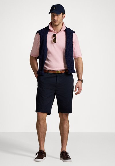 Vest bleumarin din tricotaj cu model în carouri, purtat peste un polo cu guler roz deschis, pantaloni scurți negri, curea maro și pantofi casual închiși la culoare. Accesoriile includ ochelari de soare și o șapcă.