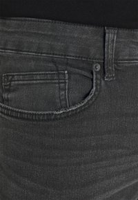 Pantalones vaqueros de mezclilla gris oscuro con un bolsillo frontal, con un diseño sencillo y una textura ligeramente deslavada. Detalle de botón metálico visible.