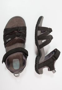 Mustat sandaalit, joissa on säädettävät velcro-nauhat, pehmeät harmaat pohjat ja teksturoitu kumipohja. Kevyt muotoilu ulkokäyttöön.