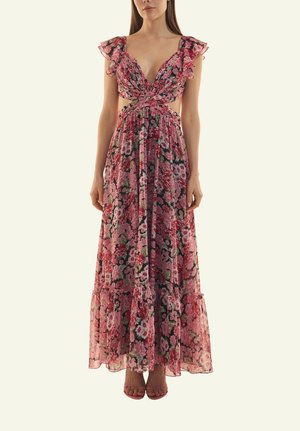 Frau trägt ein ärmelloses pinkfarbenes Maxikleid mit Blumenmuster, Rüschenschultern und Ausschnitten an der Taille, steht vor einem schlichten Hintergrund.