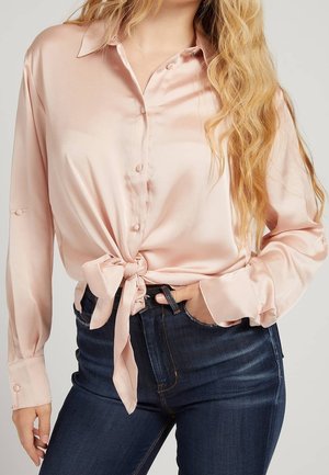 Femme portant une chemise en satin rose clair boutonnée et nouée à la taille, avec un jean taille haute bleu foncé, main dans la poche.