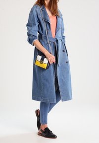 Mujer que lleva un abrigo trench de denim ceñido a la cintura, jeans ajustados azules, zapatos negros de tacón con tiras en el tobillo y sostiene un pequeño bolso colorido.