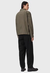 Mężczyzna z długimi włosami stoi tyłem, ubrany w oliwkową kurtkę, czarne spodnie i czarne skórzane buty na jednolitym tle.