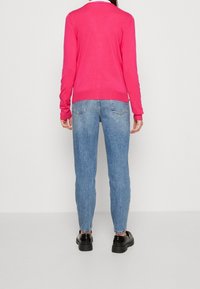 Rosa stickad cardigan med V-ringning, tillsammans med ljusblå denimjeans och svarta skor, sedd bakifrån mot en neutral bakgrund.