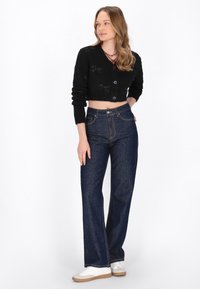 Cardigan nero corto con un tessuto a maglia testurizzato, abbinato a jeans in denim scuri a gamba larga con cuciture a contrasto e tasche anteriori. Sneakers bianche.