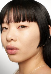 Gros plan d'un visage avec une peau lisse, des cheveux noirs raides avec une frange droite et des lèvres pulpeuses. Un arrière-plan neutre met en valeur les traits du visage.