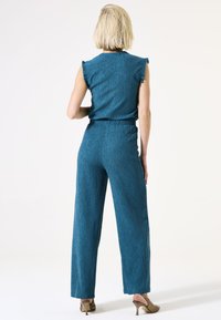 Teal jumpsuit met een gestructureerde stof, ruches op de mouwen en wijde benen. De achterkant toont een aansluitende taille en een soepele silhouet.