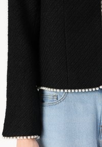 Veste noire texturée avec des détails en perles le long de l'ourlet et des poignets, portée avec un jean en denim bleu clair. Présente une coupe décontractée et un design structuré.
