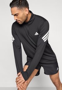 Schwarzes sportliches Langarmshirt mit hohem Kragen, weißen Drei-Streifen-Akzenten an den Ärmeln und Reißverschluss vorne; kombiniert mit schwarzen Sportshorts.
