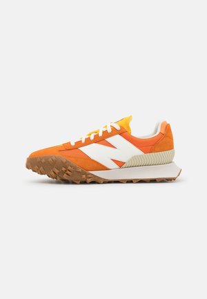 Zapatilla deportiva naranja con logo blanco "N", cordones blancos, suela marrón texturizada y talón acolchonado sobre un fondo liso y claro.
