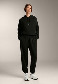 Čierna bavlnená mikina s kapucňou a sťahovacími šnúrkami, doplnená o zodpovedajúce teplákové nohavice. Oblečenie má voľný strih a elastické manžety, doplnené o biele tenisky.