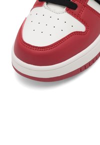 Prim-plan al părții din față a unei teneși roșii, albe și negre, cu piele albă perforată și șireți negri.