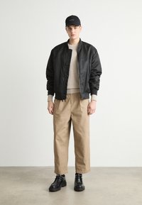 Black bomber jacket, beige sweater, and tan wide-leg pants. Black cap and ankle boots complete the outfit. Smooth textures and casual design.