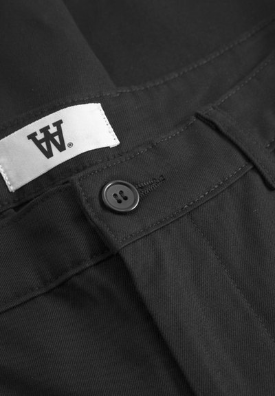 Des pantalons en tissu noir présentent une texture lisse, dotés d'un bouton noir et d'une étiquette blanche avec un marquage noir. Les coutures sont visibles le long des coutures.