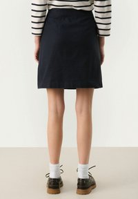 Jupe en coton bleu marine avec une coupe droite, atteignant le mi-thigh, dotée d'une texture lisse et de détails minimaux ; associée à des chaussures marron et des chaussettes blanches.