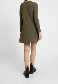 Robe verte olive à manches longues côtelées avec une coupe légèrement évasée. Portée avec des bottines noires ornées d'accents jaunes et d'une semelle épaisse.