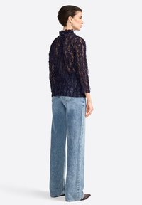 Una donna con un'acconciatura elegante a chignon indossa un top di pizzo blu navy e jeans ampi di un blu chiaro, mentre posa di lato contro uno sfondo neutro.
