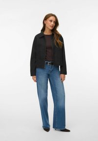 Femme portant une veste noire à fermeture éclair, un haut foncé, un jean bleu à jambes larges, une ceinture noire et des chaussures pointues foncées, se tenant avec une expression neutre.