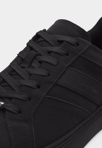 Baskets basses noires en cuir texturé, lacets plats noirs, avec le logo Calvin Klein sur le côté.