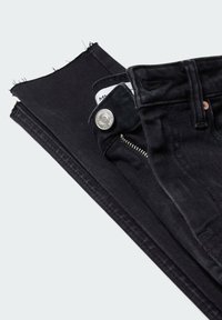 Jean noir plié avec un ourlet effiloché, bouton visible, fermeture éclair et passants de ceinture sur un fond clair.