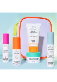 Cinq produits de soin pour la peau colorés Drunk Elephant, comprenant des sérums, une crème et un nettoyant, disposés devant une petite trousse de voyage violette et orange.