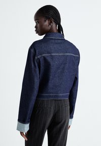 Avkortad denimjacka i mörkblått med kontrasterande ljusblå ärmar, vilket har synlig sömnad och en klassisk kragdesign.