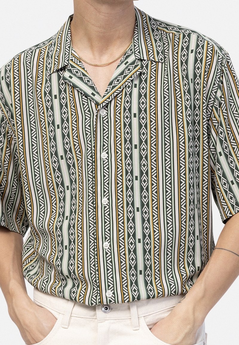 Personne portant une chemise à manches courtes à boutons, à rayures verticales géométriques vertes, blanches et dorées, et un pantalon clair, les mains dans les poches.