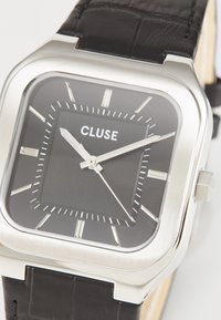 Τετράγωνο ασημένιο ρολόι από ανοξείδωτο χάλυβα με μαύρο καντράν, λευκούς δείκτες ωρών και μαύρη δερμάτινη λουρί. Φέρει το όνομα μάρκας "CLUSE."